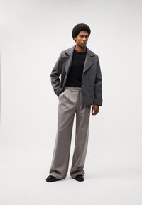 Manteau en laine gris avec boutonnière à l'avant, associé à un pantalon beige taille haute. Pull noir en dessous, complété par des chaussures noires.