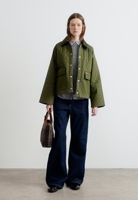 Γυναίκα που φοράει πράσινο καπιτονέ μπουφάν, ριγέ πουκάμισο, γκρι πουλόβερ, σκούρα τζιν με φαρδύ πόδι, μαύρα παπούτσια, κρατώντας καφέ καρό τσάντα, στέκεται σε απλό φόντο.