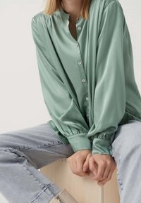 Blusa verde chiaro con bottoni, caratterizzata da maniche a sbuffo e colletto alto, realizzata in un tessuto morbido e leggero. Abbinata a jeans chiari.