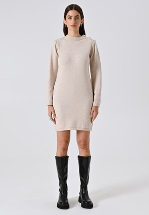 Abito lavorato a maglia beige con maniche lunghe e collo alto, caratterizzato da bottoni decorativi sulle spalle. Abbinato a stivali neri sopra il ginocchio.