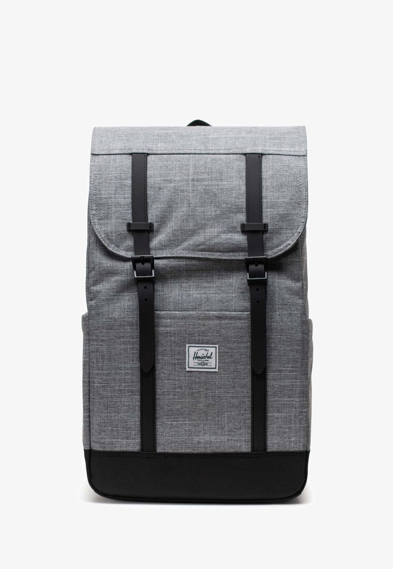 Szary plecak z tkaniny o uporządkowanej konstrukcji, czarne skórzane paski, górna klapa i wyraźna naszywka z logo na przednim panelu.
