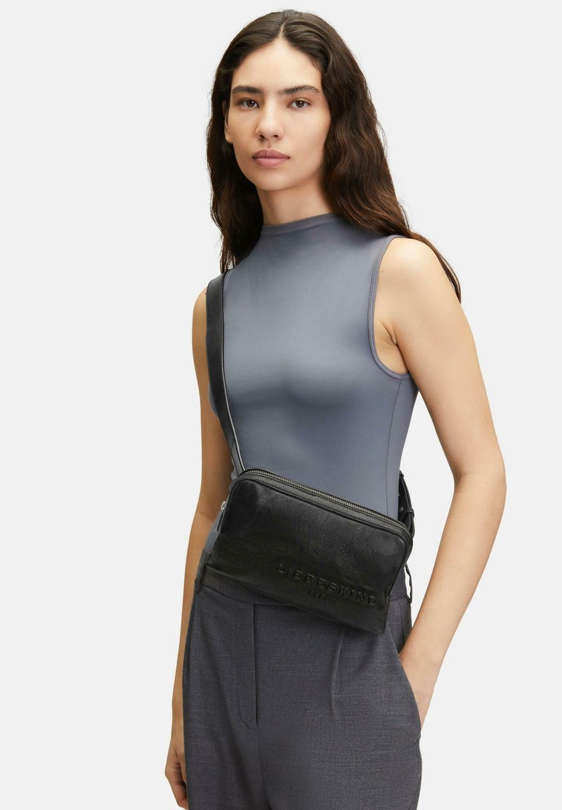 Borsa a tracolla nera in pelle con finitura texturizzata, zip argentata e logo impresso. Indossata sopra una maglietta grigia senza maniche.
