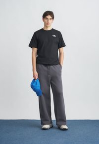 Zwart T-shirt met korte mouwen en wit logo, grijze wijde broekspijpen, en een blauwe honkbalpet die in de linkerhand wordt gehouden. De schoenen zijn lichtgrijs met donkere zolen.