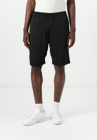 Black mesh athletic shorts with an elastic waistband and drawstring, featuring side pockets and a loose fit. Paired with white sneakers.
