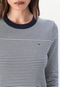 Marineblå og hvit stripete strikkegenser med rund hals og ribbestrikkede mansjetter. En liten rød logo på venstre bryst tilfører detalj.