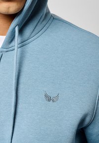 Sudadera de color azul claro fabricada con un tejido suave, con un bolsillo canguro en la parte frontal, puños acanalados y un logo bordado con alas en el pecho.