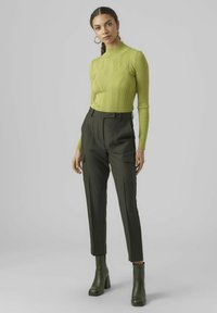 Grüner gerippter Rollkragenpullover, enganliegend. Olivgrüne Cargo-Hosen mit Seitentaschen. Massige olivfarbene Stiefeletten. Einfarbiger grauer Hintergrund.