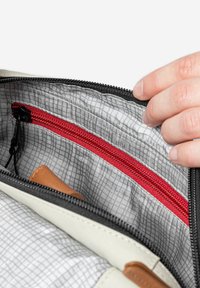 Hand opening gray fabric bag showing interior with red zipper pocket and a brown leather item inside.