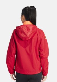 Veste rouge avec une capuche, dotée d'une doublure intérieure orange. Comprend des poignets élastiques et une coupe ample, fabriquée en un matériau léger et brillant.