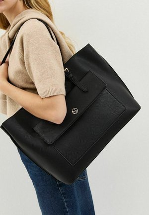 Donna che porta una grande borsa tote in pelle nera testurizzata con tasca a patta, indossa un maglione beige a maniche corte e jeans blu su sfondo neutro.