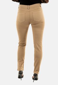 Donna che indossa pantaloni slim color cammello e tacchi alti neri, in piedi con la schiena rivolta verso la fotocamera.
