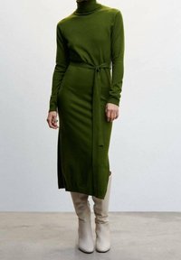 Robe en maille verte à manches longues avec un col roulé et une ceinture à la taille, dotée d'une fente latérale et associée à des bottes blanches montantes jusqu'aux genoux.