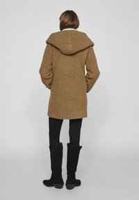 Abrigo marrón texturizado con una gran capucha, mangas largas y longitud hasta la mitad del muslo, combinado con pantalones negros ajustados y botas negras hasta la rodilla.