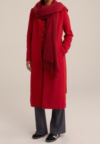 Röd ullmix lång kappa med framknappar, kombinerad med en matchande oversize-scarf. Mörkgrå byxor och svarta sneakers fulländar looken.