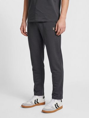 Person trägt schwarze Freizeithose und weiße Sneaker mit schwarzen Chevron-Streifen und steht vor hellem Hintergrund.