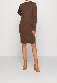 Abito a maglia marrone a maniche lunghe con collo alto, polsini a costine e silhouette aderente. Abbinato a stivaletti alla caviglia lucidi color tan e un colletto chiaro.