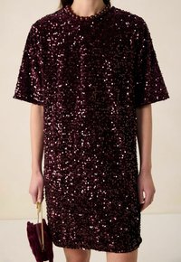 Robe en sequins bordeaux à coupe décontractée, manches courtes, col rond et texture scintillante, complétée par un collier de perles.