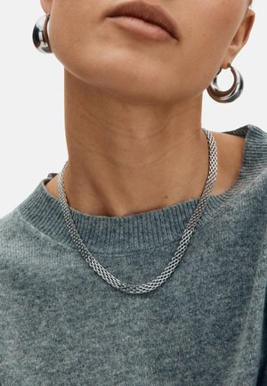 Srebrna ogrlica z verižnim vzorcem, nošena preko teksturiranega sivega puloverja. Model nosi tudi velike kovinske obročaste uhane.