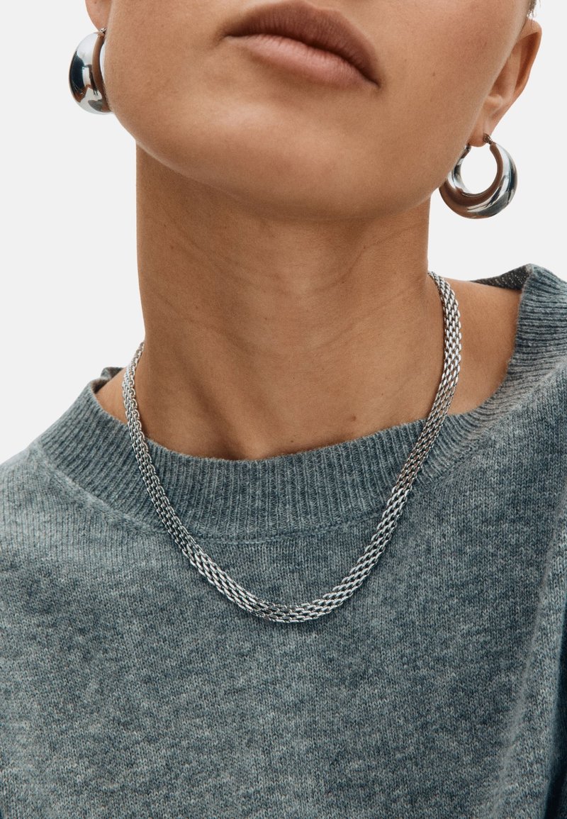 Strieborný náhrdelník s pleteným dizajnom, nosený cez textúrovaný sivý sveter. Modelka má tiež veľké kovové kruhové náušnice.