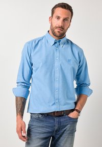 Mies, jolla on käännetyt hihat, pukeutunut vaaleansiniseen nappipaitaan, farkkuihin, ruskeaan vyöhön, kelloon ja vasemman kyynärvarren tatuointiin, seisomassa valkoista taustaa vasten.