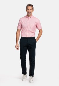 Roze overhemd met korte mouwen en een borstzak, gecombineerd met donkere broek en witte sneakers, staande op een neutrale achtergrond.