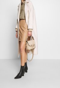 Beige leather skirt paired with an olive green shirt and a white coat. Black ankle boots, beige handbag with a textured pattern, and chain detail.