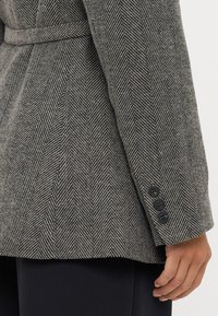 Blazer em padrão pied-de-poule em tons de cinza, com cintura cingida por um cinto, ombros estruturados e três botões escuros nos punhos.