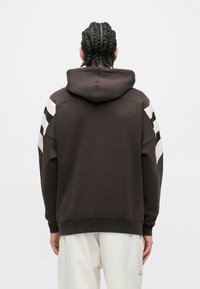 Hátulról látható személy, akinek befonott haja van, fekete kapucnis pulóvert visel fehér és fekete csíkokkal az ujjakon, valamint törtfehér nadrágot.