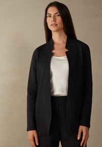 Zwarte, open voorjaarsjack met een kraagloos ontwerp, gemaakt van een gladde stof. Gecombineerd met een wit satijnen top en zwarte broek.