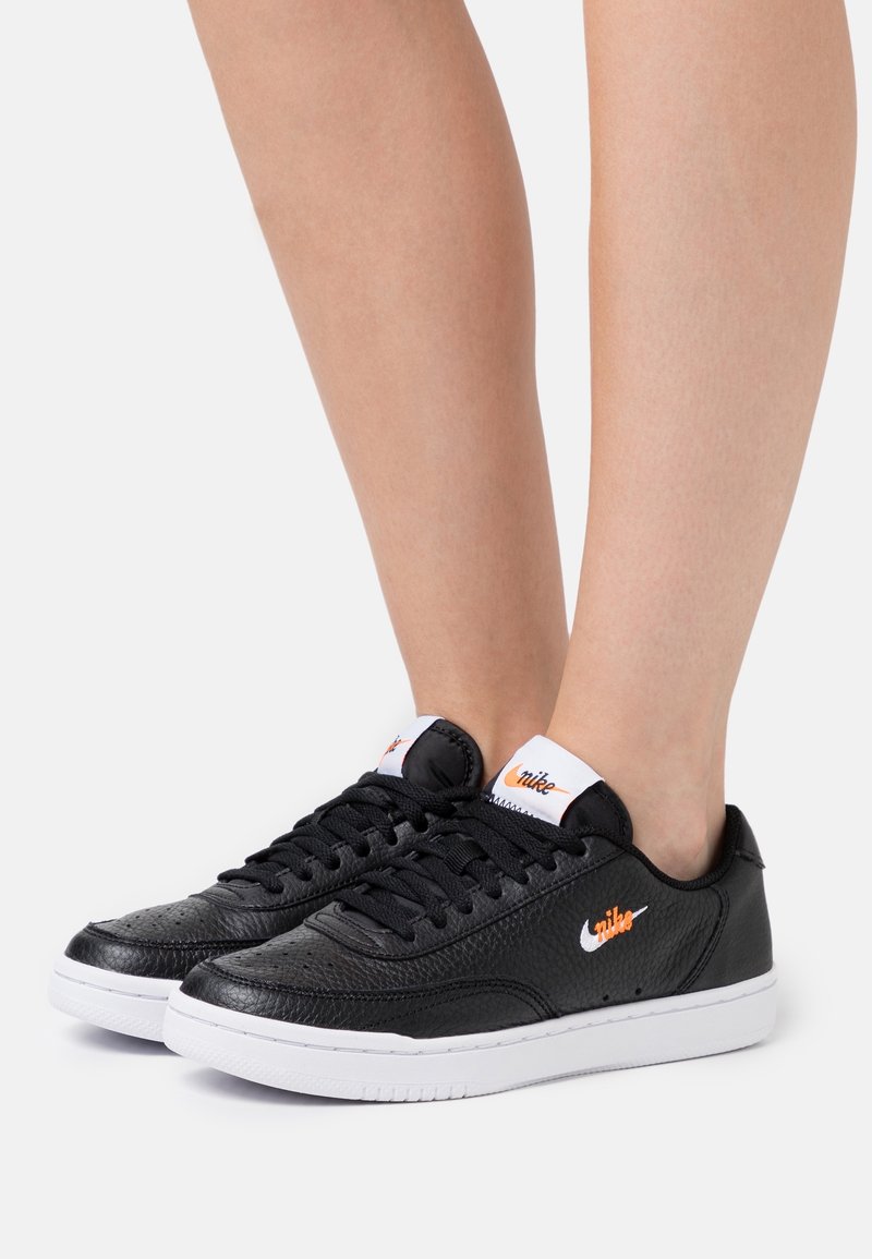 Černé tenisky Nike mají texturovaný kožený svršek, kulatou špičku, bílou podrážku a oranžové logo jako akcent na boku a jazyku.