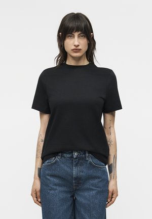 Γυναίκα με σκούρα μαλλιά που φορεί μαύρο κοντομάνικο μπλουζάκι και μπλε τζιν, στέκεται μπροστά σε απλό λευκό φόντο.