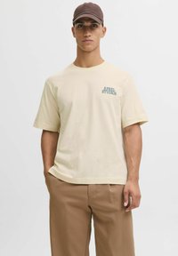 Tricou crem cu mânecă scurtă, pe care este imprimat "APREL STUDIOS" cu albastru pe piept, asortat cu pantaloni largi de culoare maro deschis.