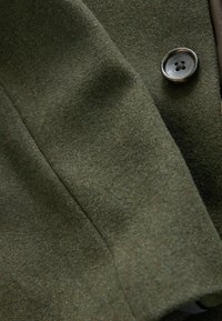 Olivgrüner Wollstoff mit einer sanften Textur, der einen glatten, glänzenden Metallknopf und dezente Nähdetails aufweist.