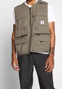 Khaki nytta väst med flera fickor, dragkedjor och detalj i form av en ring. Tillverkad i slitstarkt tyg; har dragkedja fram och en avslappnad passform.