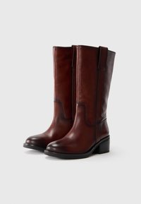 Botas de cuero marrón hasta la rodilla con una textura suave, punta redondeada y un tacón bajo en bloque. Presentan un diseño de costura contrastante a lo largo del lado.
