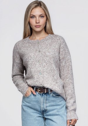 Une femme présente un cardigan en maille gris clair boutonné, associé à un jean bleu et à une ceinture marron, sur un fond uni.