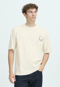 Cremefarvet bomulds T-shirt med rund halsudskæring, korte ærmer og et mørkt grafisk design på venstre brystområde.