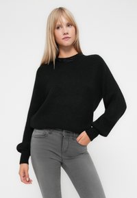 Pull en maille noire avec un col rond et des manches longues, associé à un jean gris ajusté. Le pull présente un motif texturé au niveau du col.