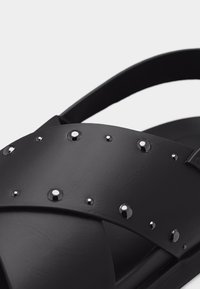 Μαύρο σανδάλι με φαρδιές διασταυρούμενες λουράκια διακοσμημένα με γυαλιστερές μαύρες στρογγυλές λεπτομέρειες και λουρί που αγκαλιάζει την φτέρνα.