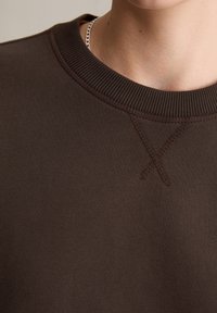 Brązowa bluza z prążkowanym dekoltem typu crew, teksturowaną tkaniną i ozdobnym przeszyciem w kształcie litery 'X' w pobliżu kołnierza.