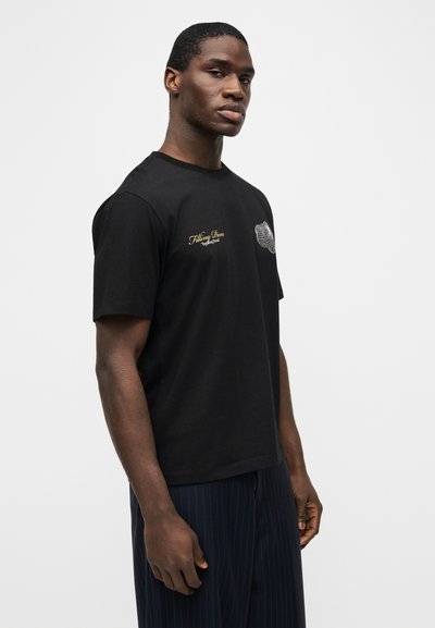 Νεαρός άνδρας με κοντά μαλλιά που φοράει μαύρο γραφιστικό μπλουζάκι και σκούρο παντελόνι με λεπτές ρίγες, στέκεται μπροστά σε ένα απλό φωτεινό φόντο.