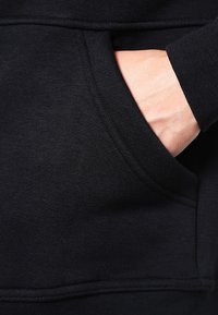 Svart hoodie med en framficka i kangurustil, mjuk textur och ribbade ärmslut. Sömnadsdetaljer syns längs sömmarna.