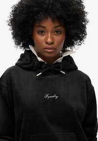 Felpa in corduroy nera con coulisse, dotata di tasca anteriore e logo "Superdry" ricamato in bianco. Il tessuto appare morbido e testurizzato.