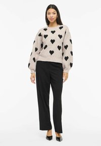 Jemně růžový svetr s černými srdíčkovými vzory, dlouhými nafouklými rukávy a zaobleným výstřihem, doplněný o černé kalhoty s rovnými nohavicemi a špičaté boty.