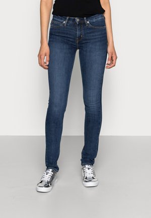 Personne portant un jean skinny bleu foncé et des baskets argentées métalliques, debout devant un fond clair uni.