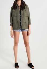 Olivgrön arbetsjacka med knäppning, med två bröstfickor och uppvikt ärmar, tillsammans med jeanstashorts och svarta sandaler.