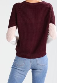 Frau trägt einen gerippten Strickpullover in Burgunderrot mit hellrosa und weißen Blockärmeln, kombiniert mit hellblauen Jeans.
