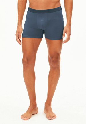 Modello maschile che indossa boxer aderenti blu scuro, in piedi a piedi nudi su uno sfondo bianco uniforme.