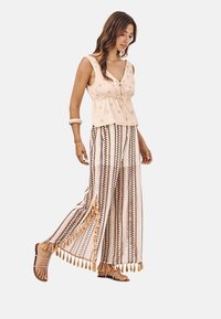 Top sin mangas de color beige claro con un detalle de lazo, combinado con pantalones de pierna ancha de estampado que presentan colores terrenales y acentos de borlas. Tejido suave.