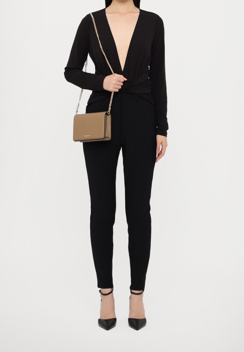 Femme portant un haut noir à manches longues avec un décolleté en V profond, un pantalon noir moulant, des talons noirs avec bride à la cheville et portant un petit sac marron en bandoulière.